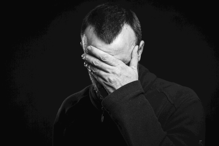 Mann mit Geheimratsecken hält sich traurig eine Hand vor das Gesicht vor schwarzem Hintergrund