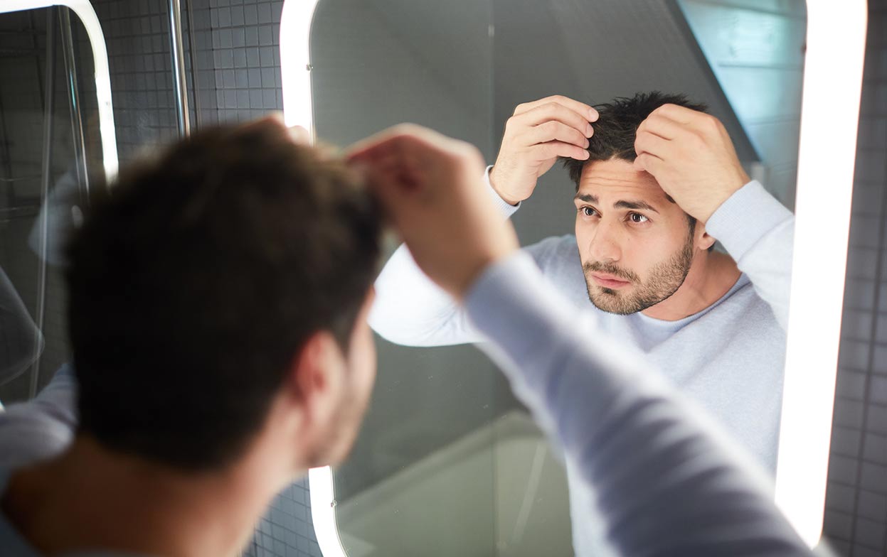 Mann steht vor Spiegel und denkt über Verhalten nach der Haartransplantation nach