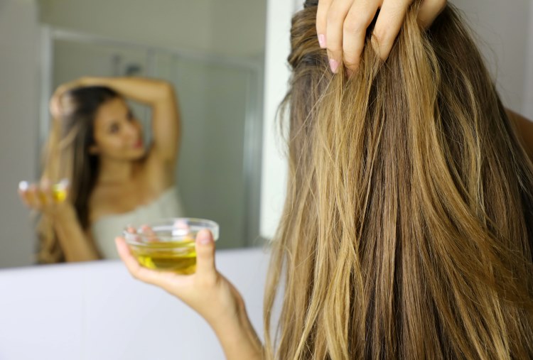 Frau behandelt Ihre Haare mit Olivenöl aus einem Schälchen und betrachtet sich im Spiegel