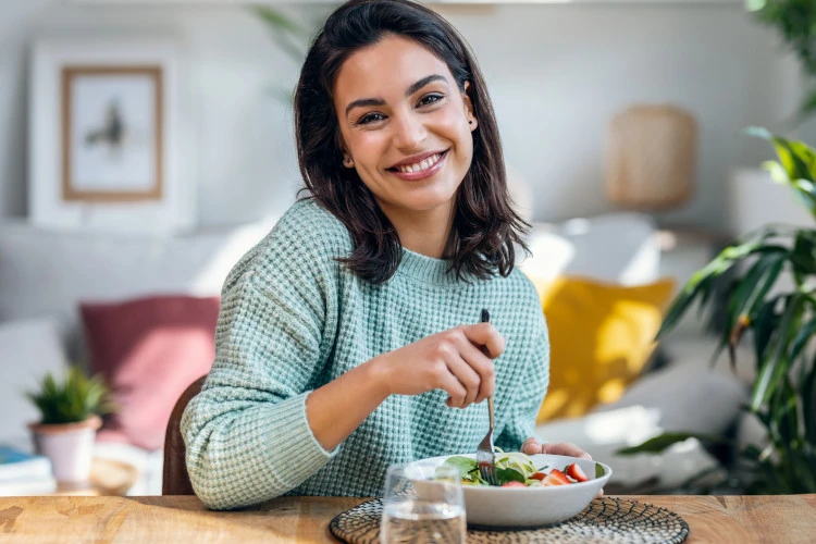 Lächelnde Frau in hellem Raum, die einen frischen Salat genießt, symbolisiert gesunde Ernährung und bewussten Lebensstil