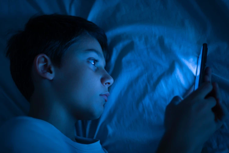 Ein Junge liegt im Bett und schaut konzentriert auf sein Smartphone, das ein blaues Licht ausstrahlt