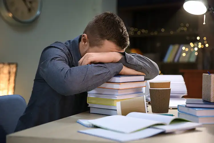 Junger Mann mit gesenktem Kopf auf einem Stapel Bücher – zeigt Überforderung und Lernstress
