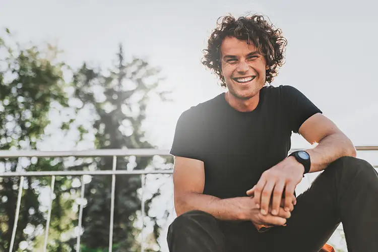 Ein lachender Mann mit lockigem Haar in sonniger Umgebung – vermittelt Lebensfreude, Zufriedenheit und ein positives Selbstbild.
