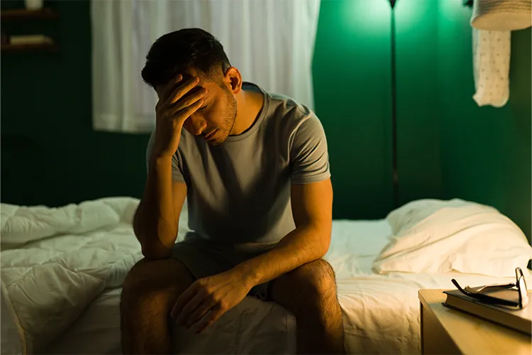 Ein Mann sitzt nachdenklich auf dem Bett, stützt den Kopf mit der Hand – symbolisiert emotionale Belastung.
