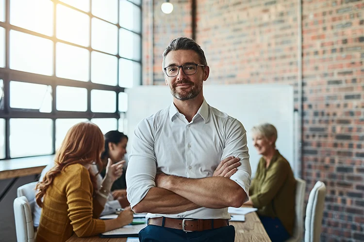 Geschäftsmann mit Brille steht mit verschränkten Armen vor einem Team im Besprechungsraum